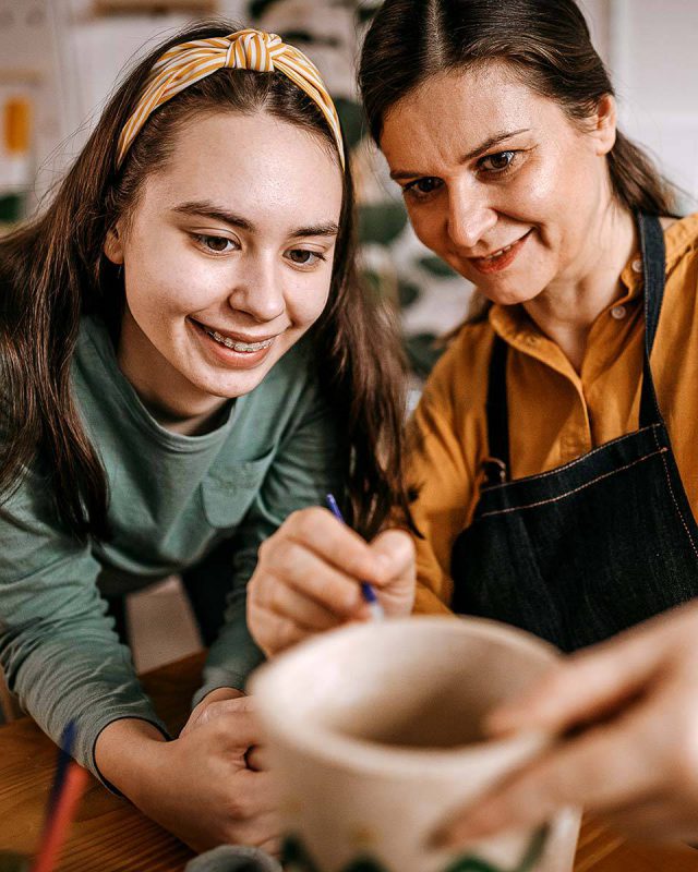 Older woman and young woman working together to paint a pottery piece they made together and smilng