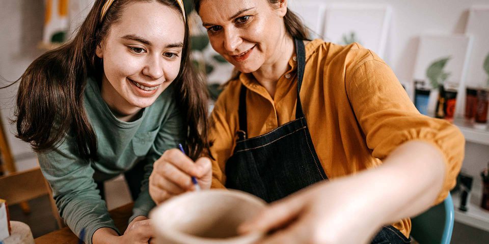 Woman and younger girl working together to shape wet clay on a pottery wheel