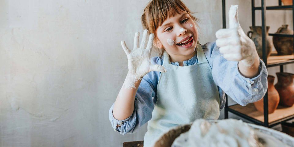 Young girl working with wet clay on a potters wheel and giving thumbs-up with clay on her face