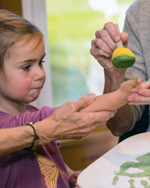 Young girl in a purple shirt having paint dotted on her hand to make a handprint on a customized clay pottery plate