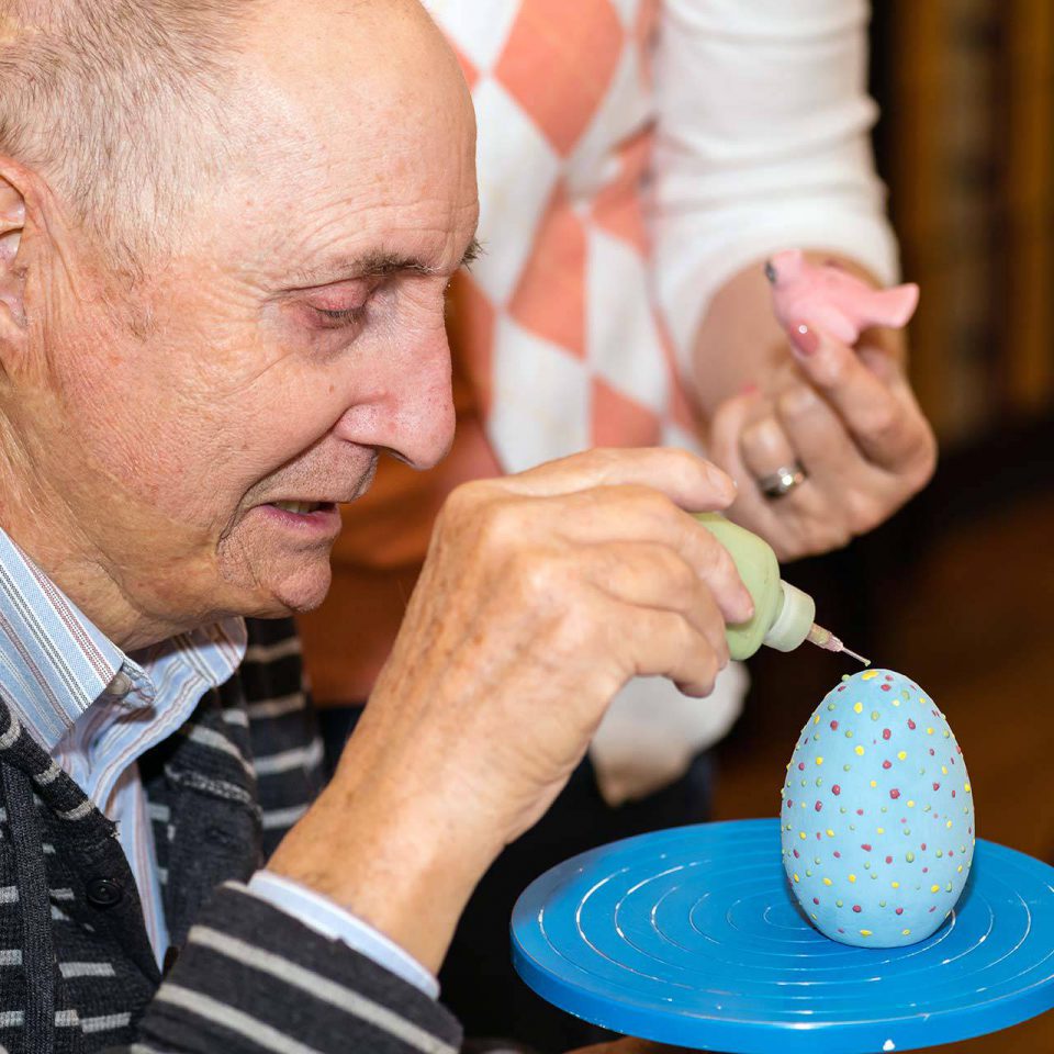 Older man using a paint bottle to make speckles and dots on a blue clay pottery egg