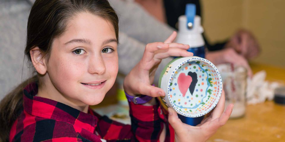 Girl wearing buffalo-check shirt holding up the pottery bowl she painted