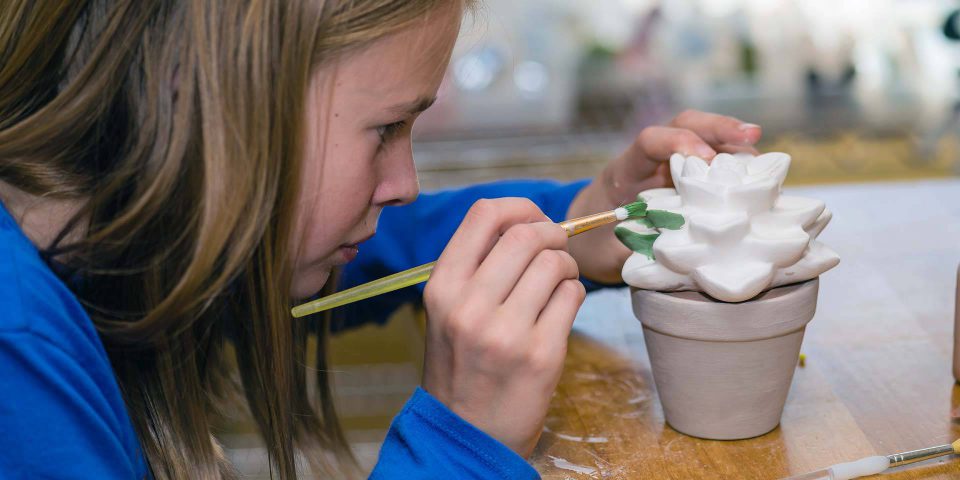 child with long brown hair and a blue long-sleeved shirt concentrating on painting a pottery succulent in a pot