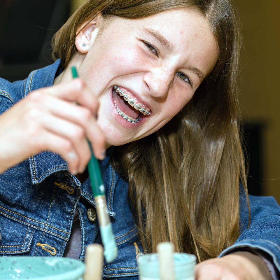 teenager with braces smiling as she uses a paintbrush to customize pottery